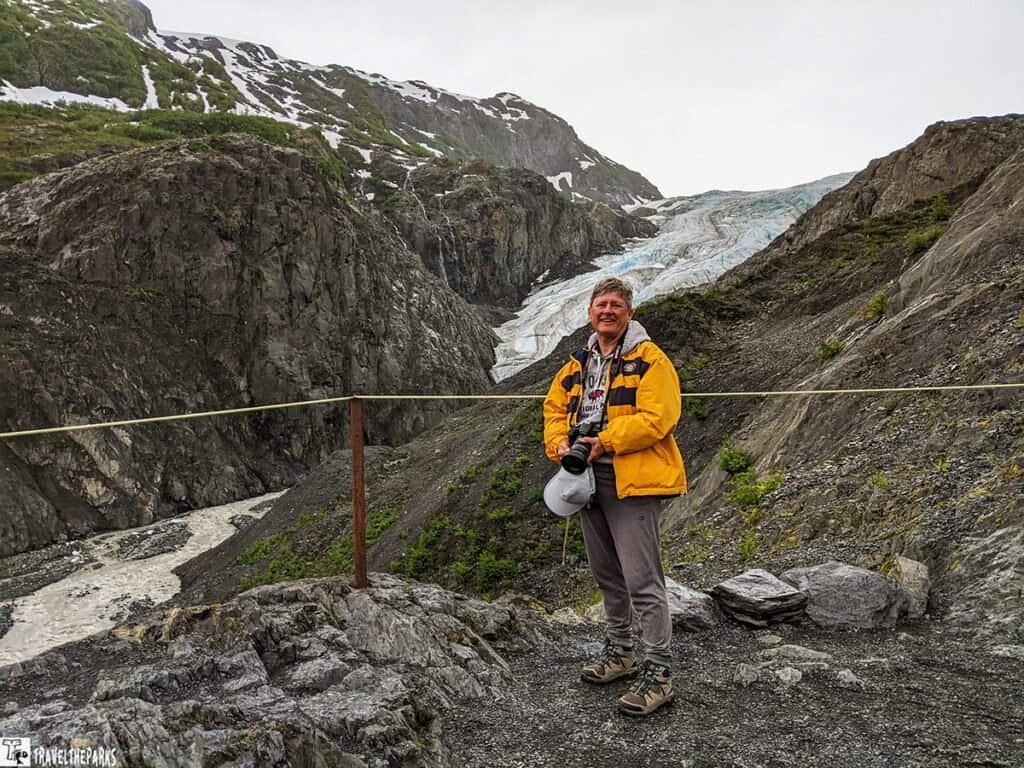 A person in a yellow jacket stands on rocky terrain with Exit Glacier in the background, holding a camera.