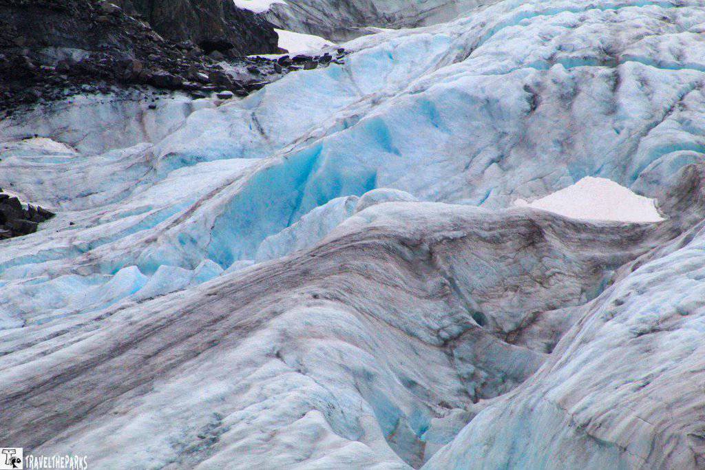 Close-up view of a glacier with blue and white ice, featuring dark streaks and rocky border.