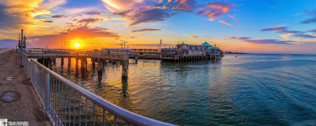 Panoramic view of Cedar Key waterfront at sunset, featuring a pier, buildings with blue rooftops, and calm waters.