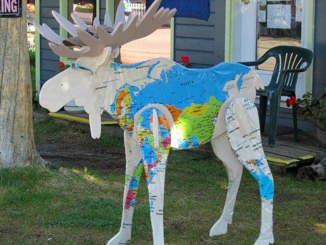 Sculpture of a moose decorated with a world map, standing on grass with a building in the background.