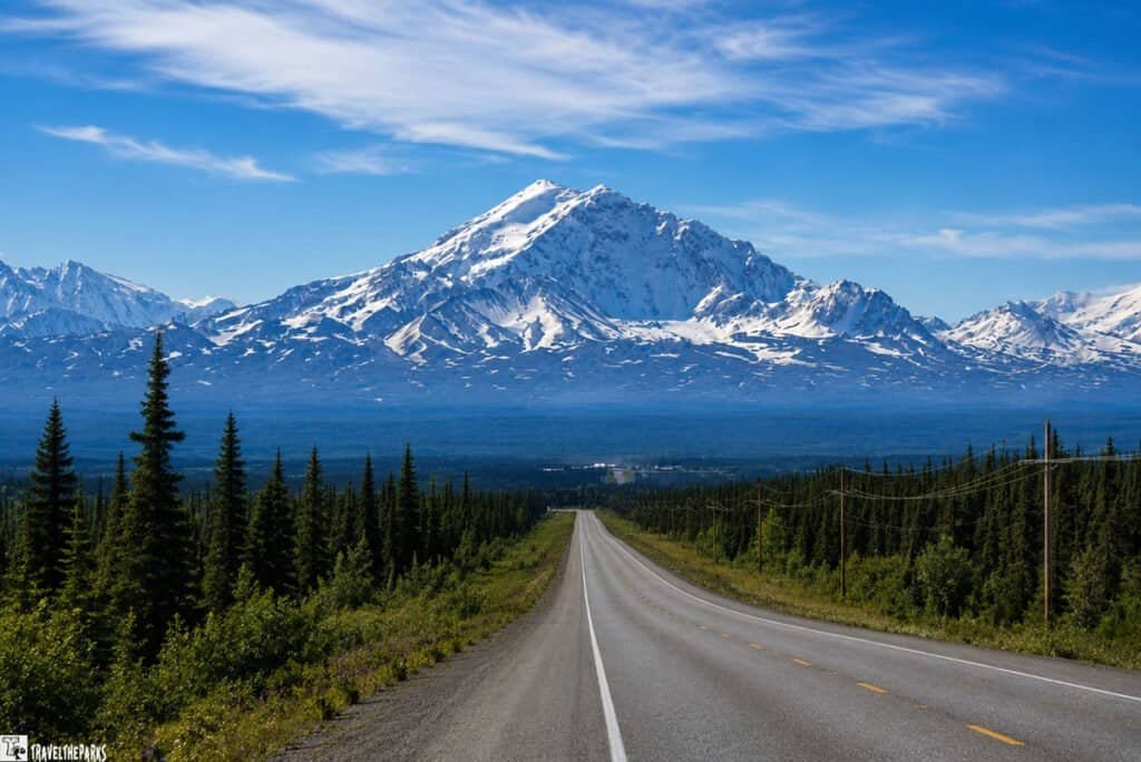 A scenic view of Mount Drum in Glenallen, Alaska, featuring a winding road leading to the mountains, which are partially covered in snow against a clear blue sky.