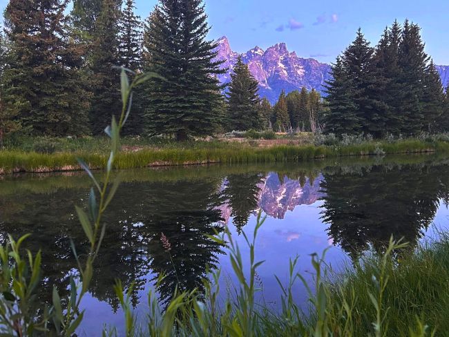 Sunset at Grand Teton with mountains reflected in a calm body of water, surrounded by trees and grass.