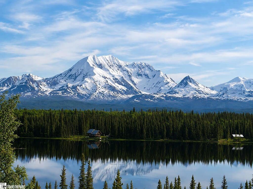View of Mt. Drum Wrangell St. Elias NP: A scenic view of a snowy mountain range reflected in a calm lake, surrounded by lush green trees and a clear blue sky with light clouds.