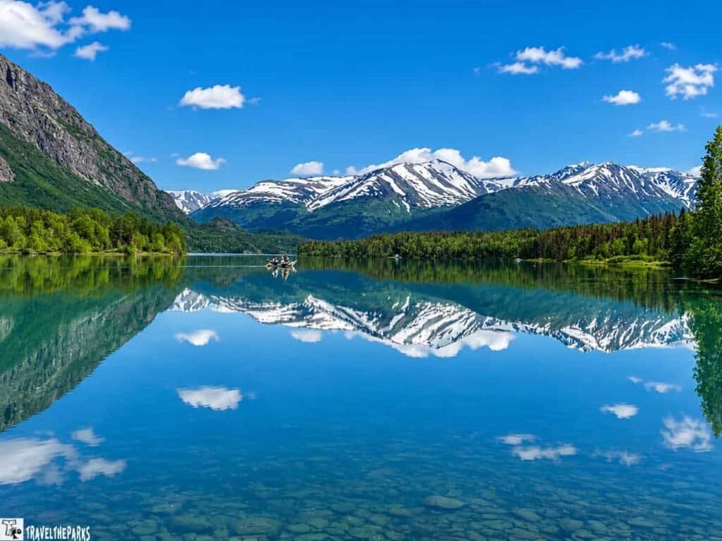 A picturesque view of the Kenai River with snow-capped mountains in the background, lush green trees along the banks, and a small boat with two people gently rowing on the mirrored water.
