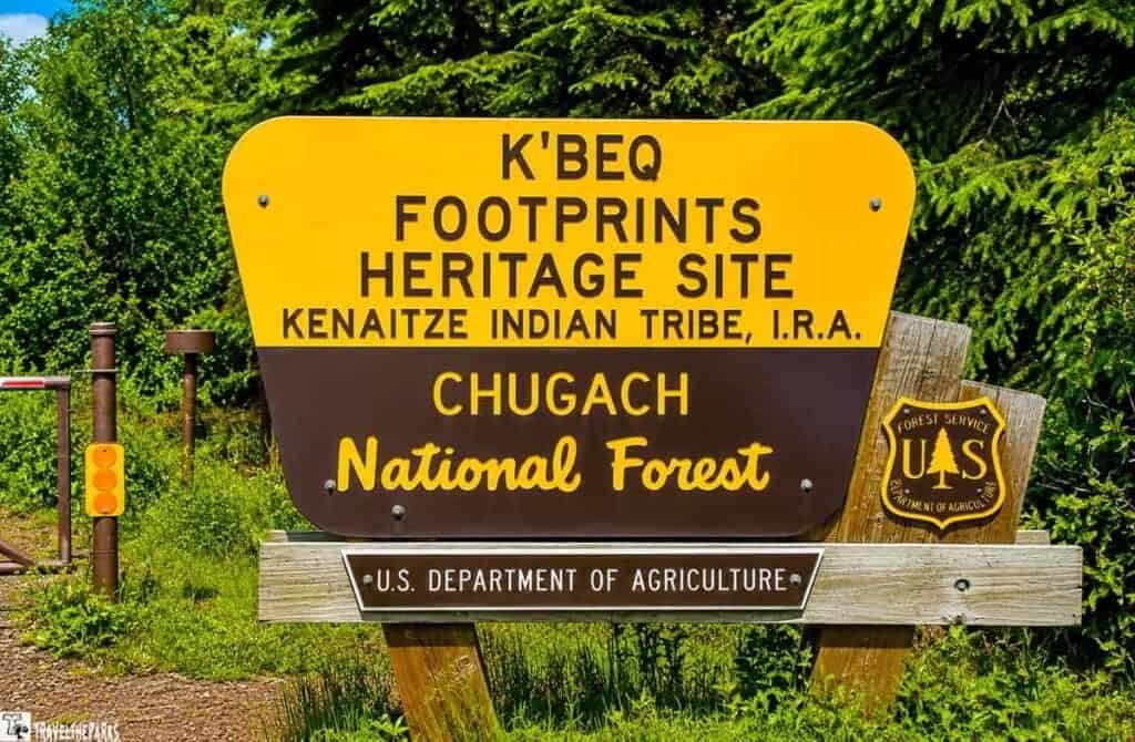 Sign reading "K'BEQ Footprints Heritage Site, Kenaitze Indian Tribe, I.R.A., Chugach National Forest" with lush greenery in the background.