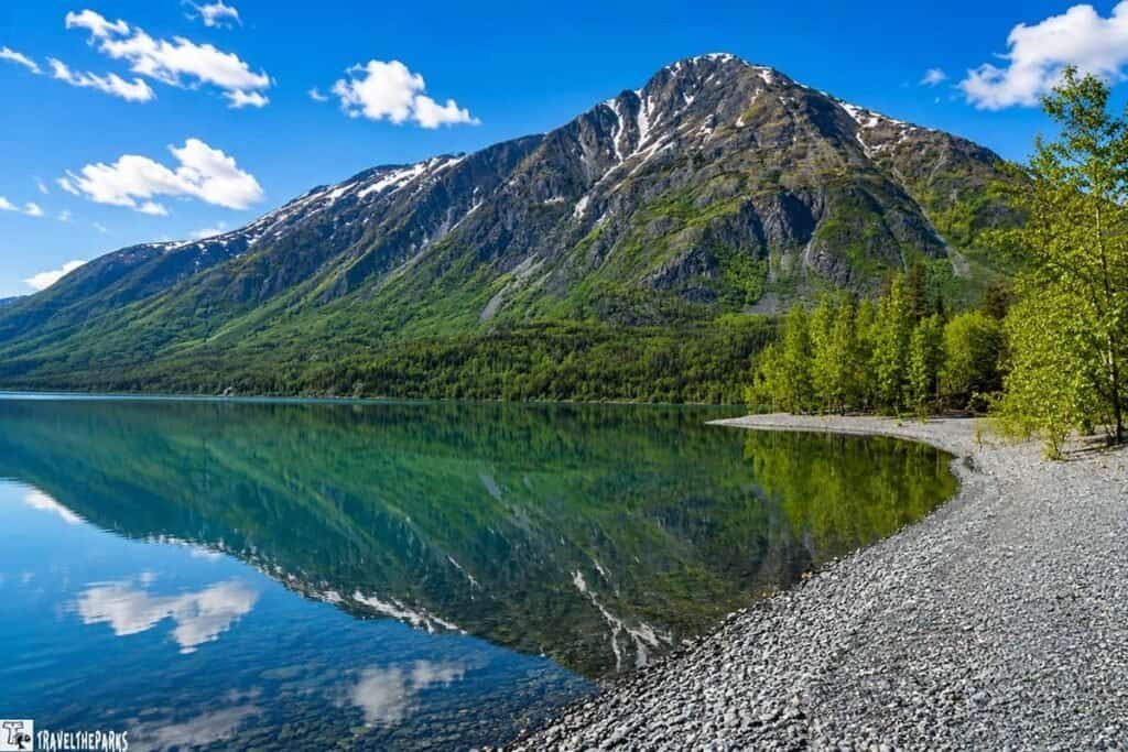 A calm lake reflects green mountains and blue skies, with a pebbly beach in the foreground and trees to the right.