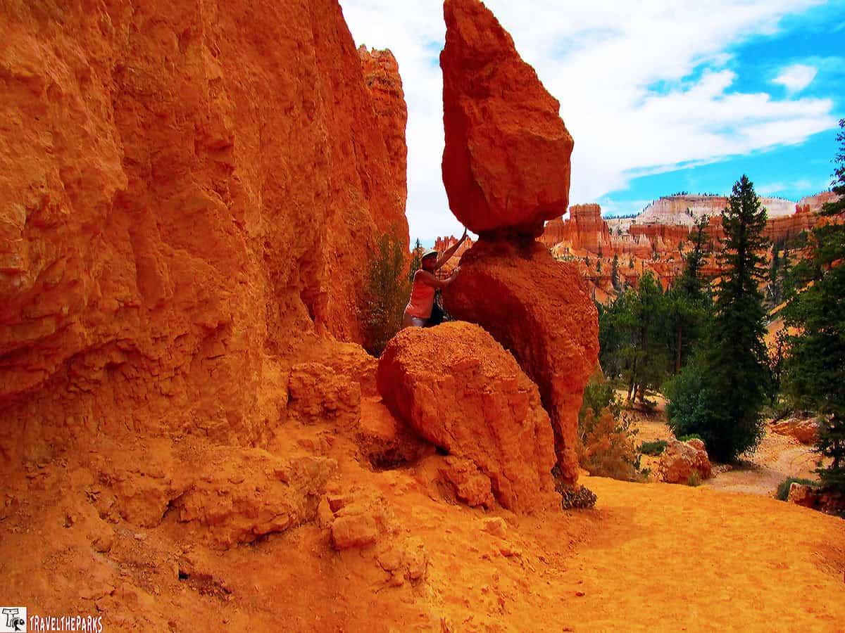 A person standing on a rock formation in Bryce Canyon, reaching toward a tall, balanced rock with a blue sky and orange hoodoos in the background.

