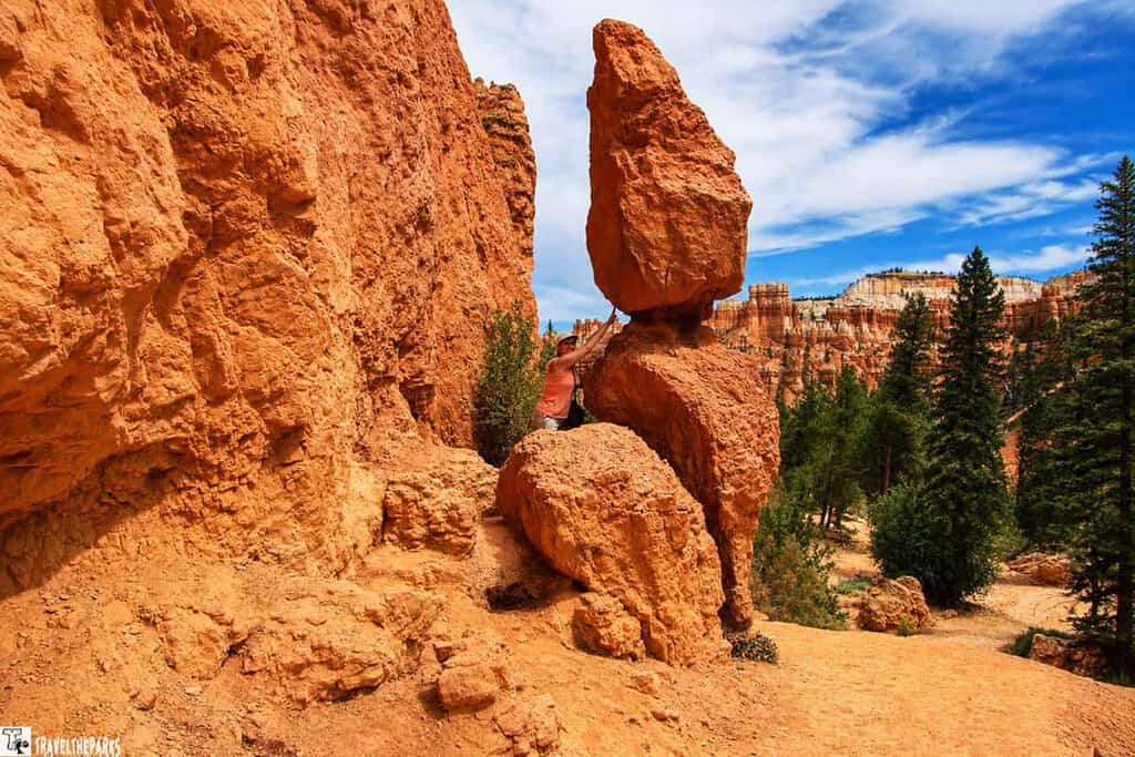 A person standing on a rock formation in Bryce Canyon, reaching toward a tall, balanced rock with a blue sky and orange hoodoos in the background.