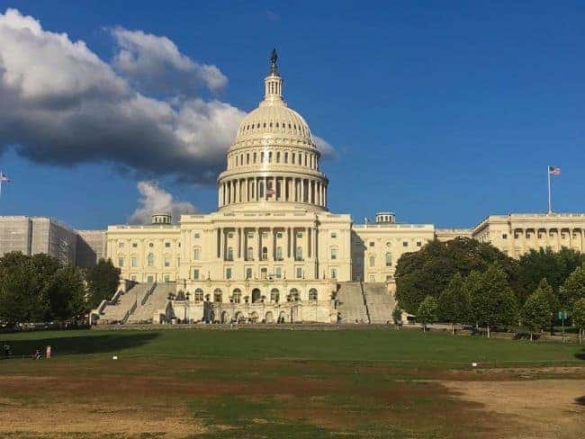 U.S. Capitol building with dome, staircase, surrounding trees, and an American flag.
