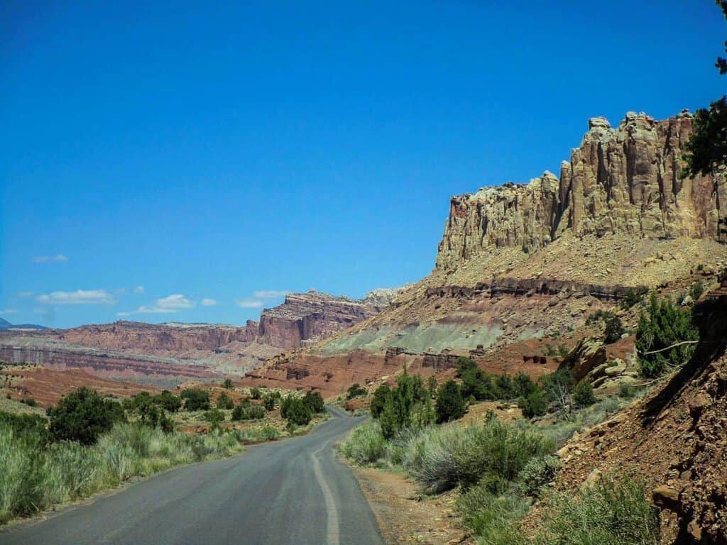 A paved road leading through a desert landscape with rocky cliffs under a bright blue sky in Capitol Reef National Park.