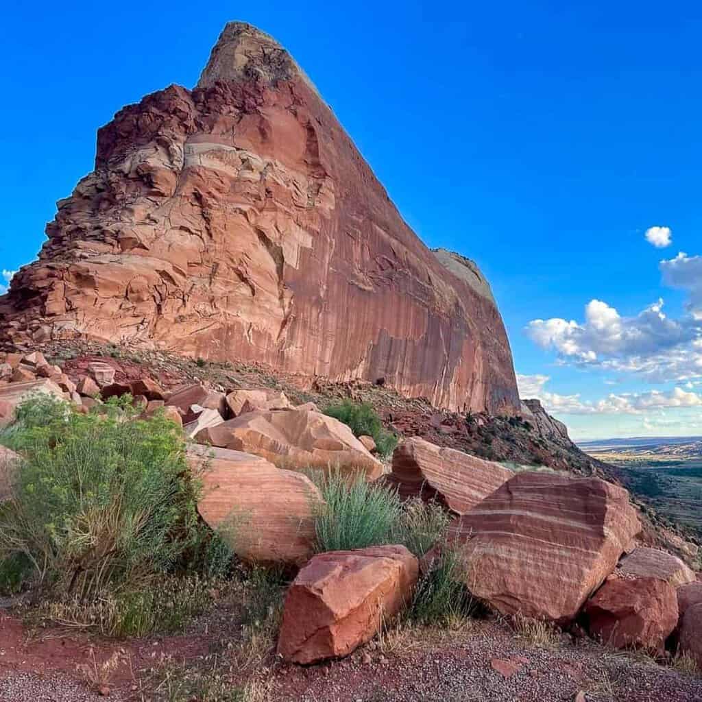 Steep red rock formation in Mule Canyon with boulders and shrubs, set against a blue sky.