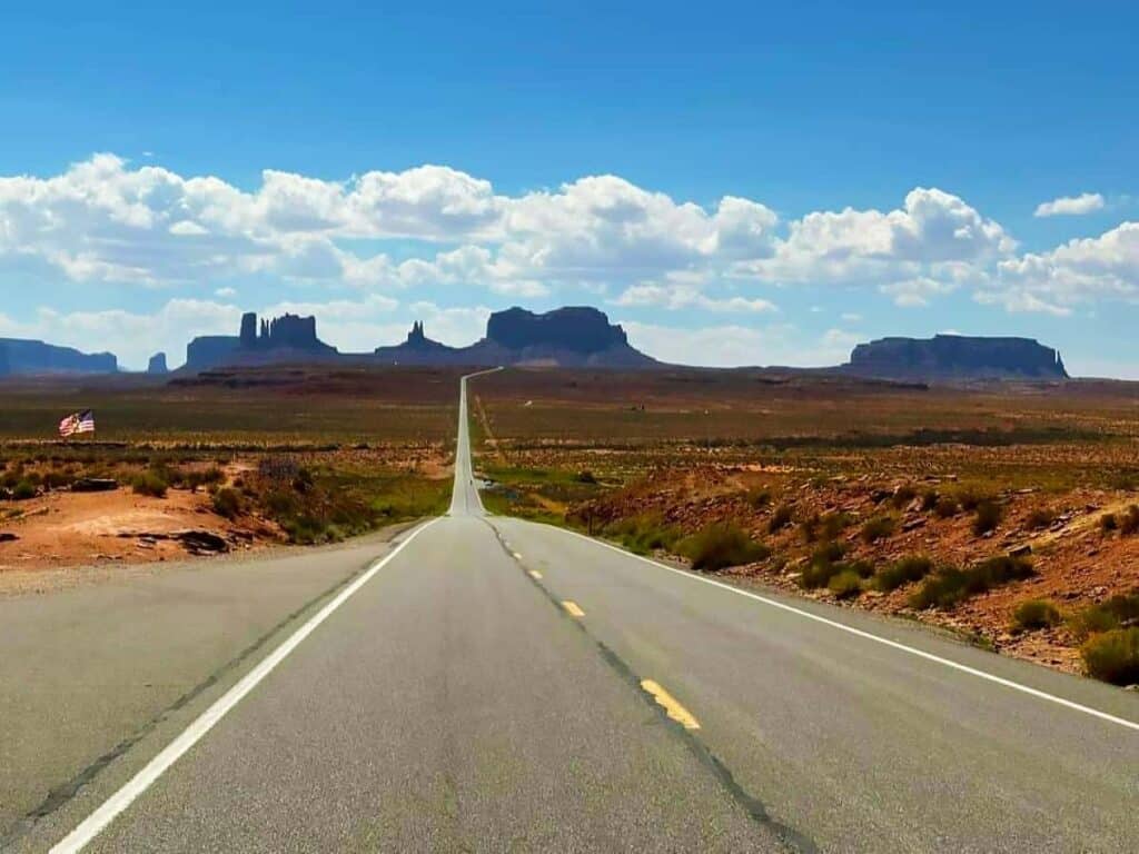 Forrest Gump Point Hwy 163: A straight road leading through a desert landscape with large rock formations in the background under a clear blue sky.