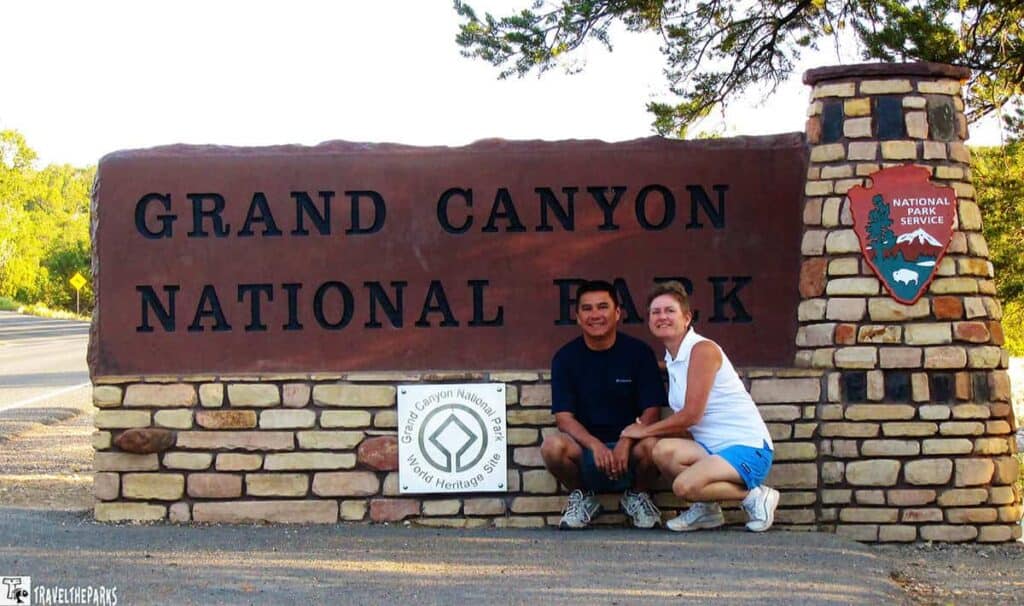 A smiling couple poses in front of the Grand Canyon National Park sign made of stone. Bright daylight enhances the scene's welcoming and adventurous feel.