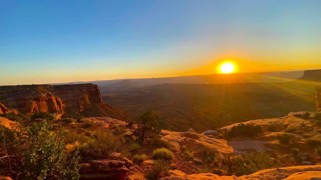 Sunrise over Canyonlands National Park, with a glowing sun over rocky cliffs and desert landscape.

