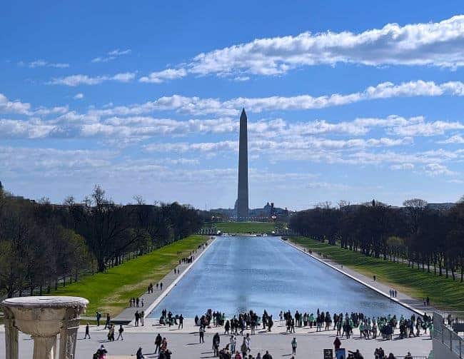 the Washington Monument, a tall, pointed obelisk standing at the center of the frame. In the foreground, the Lincoln Memorial Reflecting Pool stretches towards the monument, reflecting the sky and surrounding trees. The pathway on either side of the pool is populated with numerous people walking and gathering. The background features a clear blue sky with scattered clouds, casting soft shadows over the well-manicured green lawns and leafless trees flanking the pool.