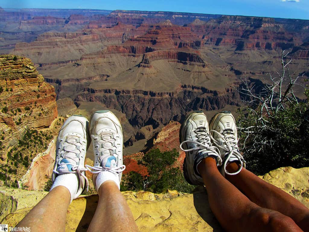 Two pairs of feet in sneakers overhanging the edge of the Grand Canyon with a view of its vast rock formations.