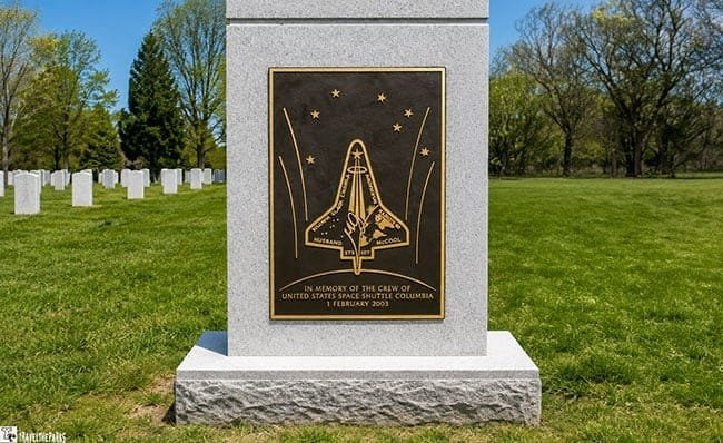 A gray stone memorial with a bronze plaque commemorating the crew of the Space Shuttle Columbia, surrounded by white gravestones in a grassy area.