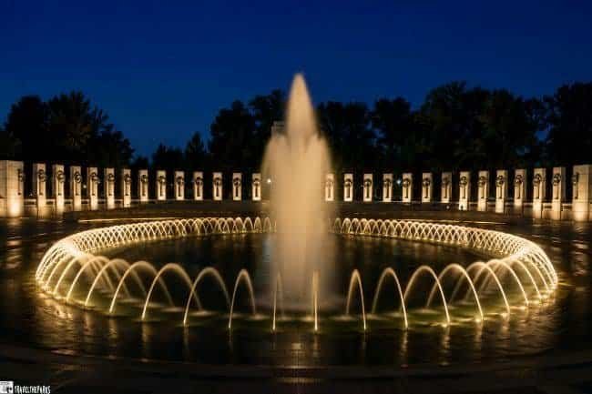 World War II Memorial-A circular illuminated fountain at night with arched water jets and background columns.
