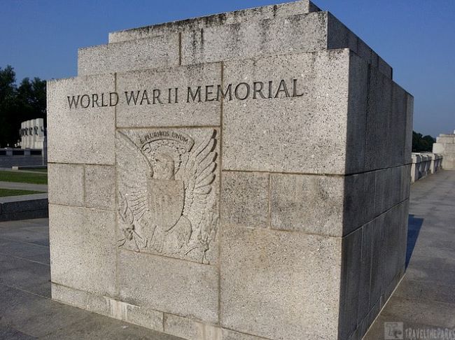 a large, rectangular stone structure at the World War II Memorial. The front face of the structure is prominently etched with the words "WORLD WAR II MEMORIAL" at the top. Below the inscription is an elaborate carving of an eagle with its wings spread, clutching a shield and surrounded by olive branches. The background of the memorial is a smooth, gray stone, with distinct block lines visible. To the left, blurred outlines of additional monument features and green trees can be seen, under a clear blue sky.