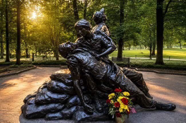 Vietnam Women’s Memorial bronze statue of a nurse holding a wounded soldier with another soldier kneeling behind her; flowers in foreground.