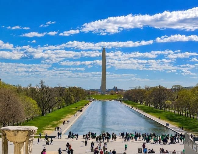 a large, rectangular stone structure at the World War II Memorial. The front face of the structure is prominently etched with the words "WORLD WAR II MEMORIAL" at the top. Below the inscription is an elaborate carving of an eagle with its wings spread, clutching a shield and surrounded by olive branches. The background of the memorial is a smooth, gray stone, with distinct block lines visible. To the left, blurred outlines of additional monument features and green trees can be seen, under a clear blue sky.
