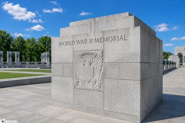a large, rectangular stone structure at the World War II Memorial. The front face of the structure is prominently etched with the words "WORLD WAR II MEMORIAL" at the top. Below the inscription is an elaborate carving of an eagle with its wings spread, clutching a shield and surrounded by olive branches. The background of the memorial is a smooth, gray stone, with distinct block lines visible. To the left, blurred outlines of additional monument features and green trees can be seen, under a clear blue sky.