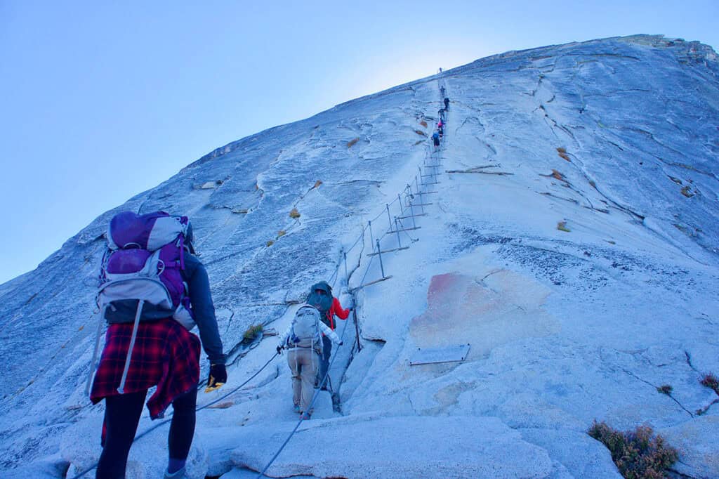 Hikers climbing the granite surface of Half Dome using metal cables in Yosemite National park.