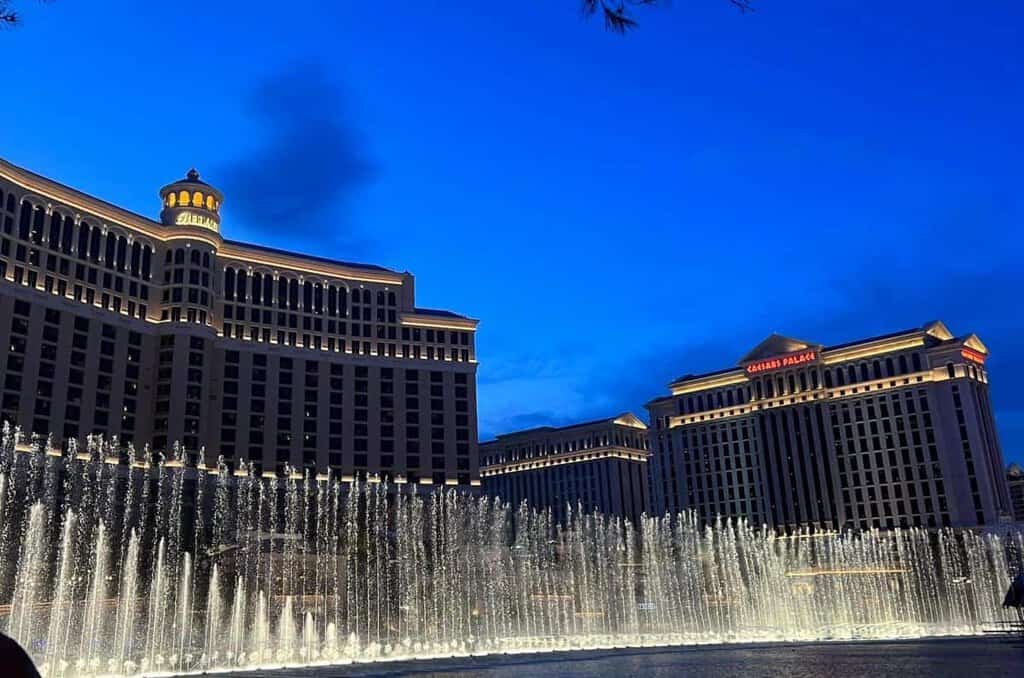 Bellagio Hotel and illuminated fountains at night with Caesars Palace visible.

