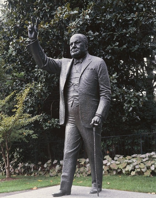 Bronze statue of Winston Churchill in a suit, holding a walking stick with his left hand and making a V-sign with his right hand, surrounded by lush greenery.