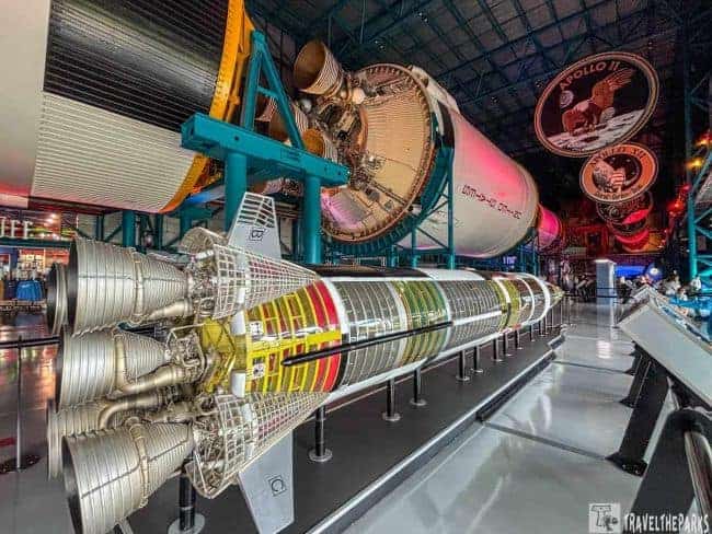 Saturn V rocket display in an exhibition hall with engine clusters and colorful segmented sections.