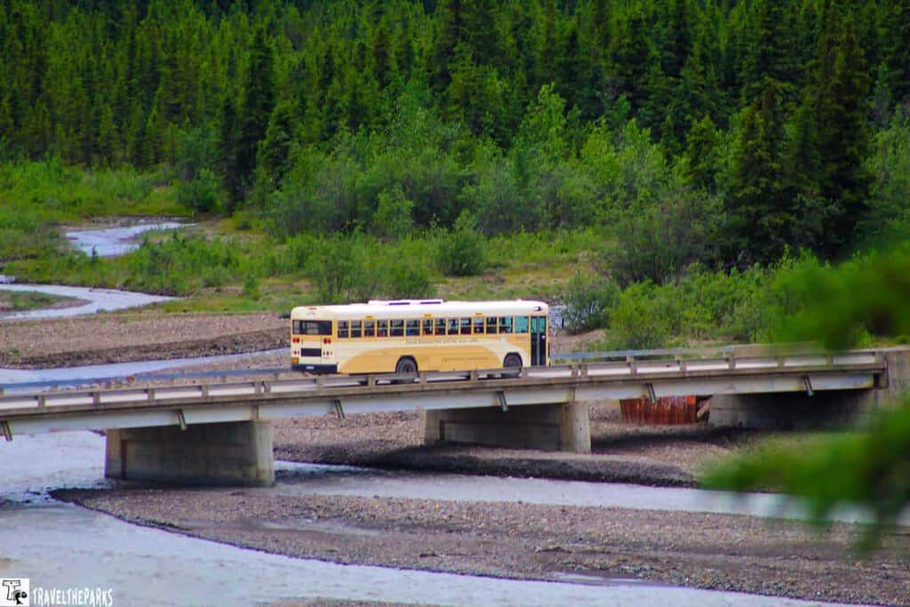 A yellow tan bus crosses the Teklankika concrete bridge over a river in Denali National Park, with a forested background.