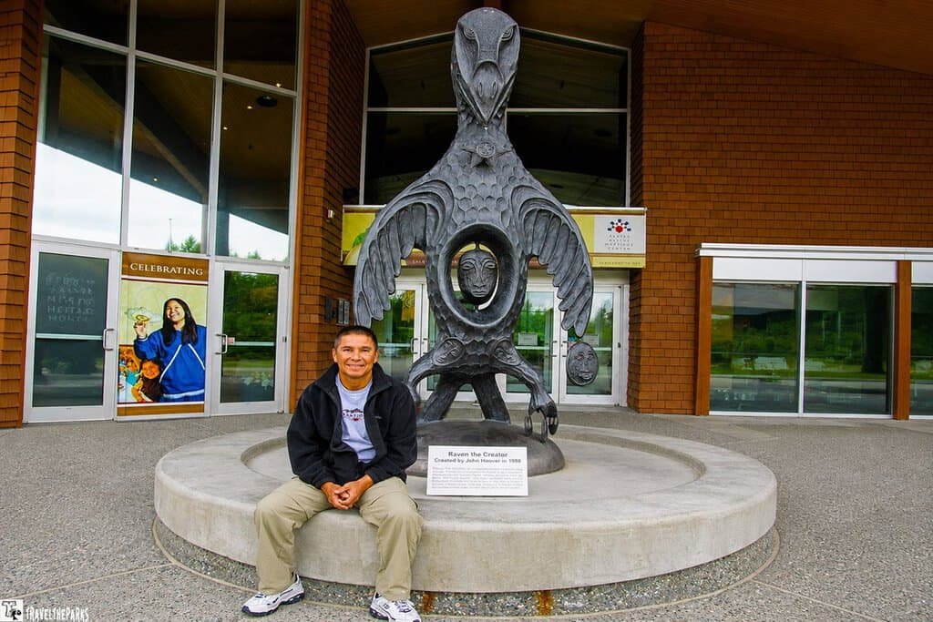 Large sculpture of "Raven the Creator" with a person sitting in front of it at the Alaska Native Heritage Center. Transcribed Text: Raven the Creator Created by Jim Schoppert, 1998 Property of Alaska Native Heritage Center Collection