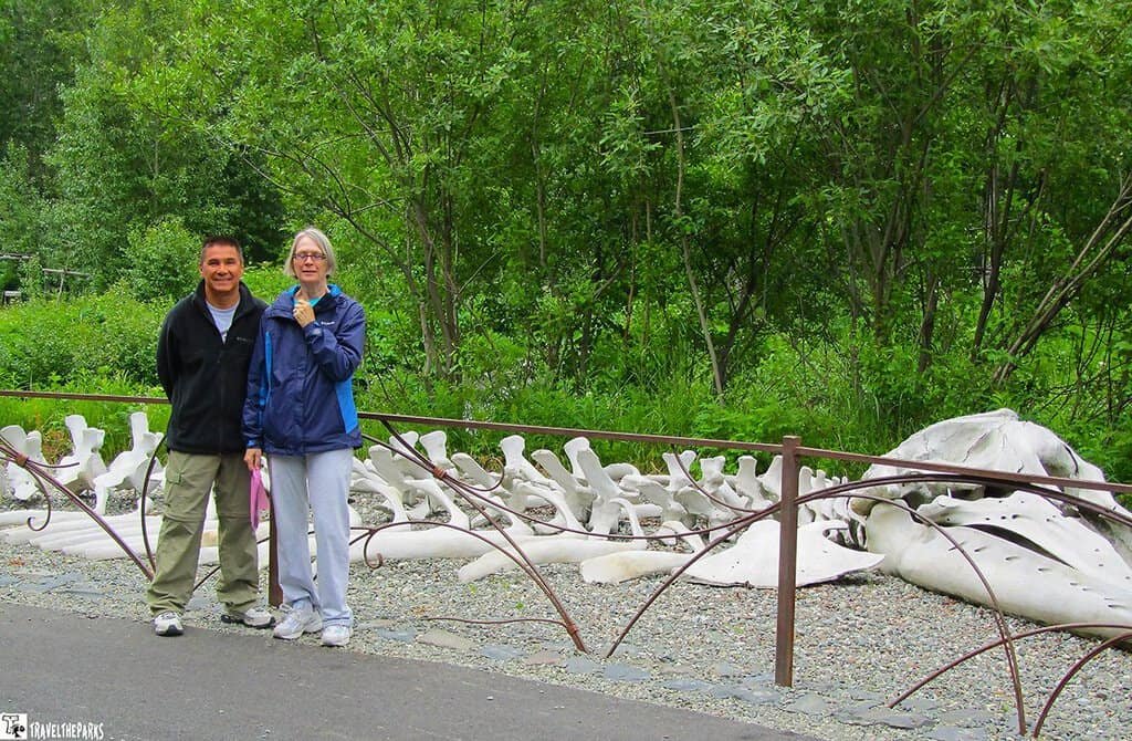 At the Alaska Heritage Center Village-Two people standing near large white bones, possibly whale bones, with a green foliage backdrop.