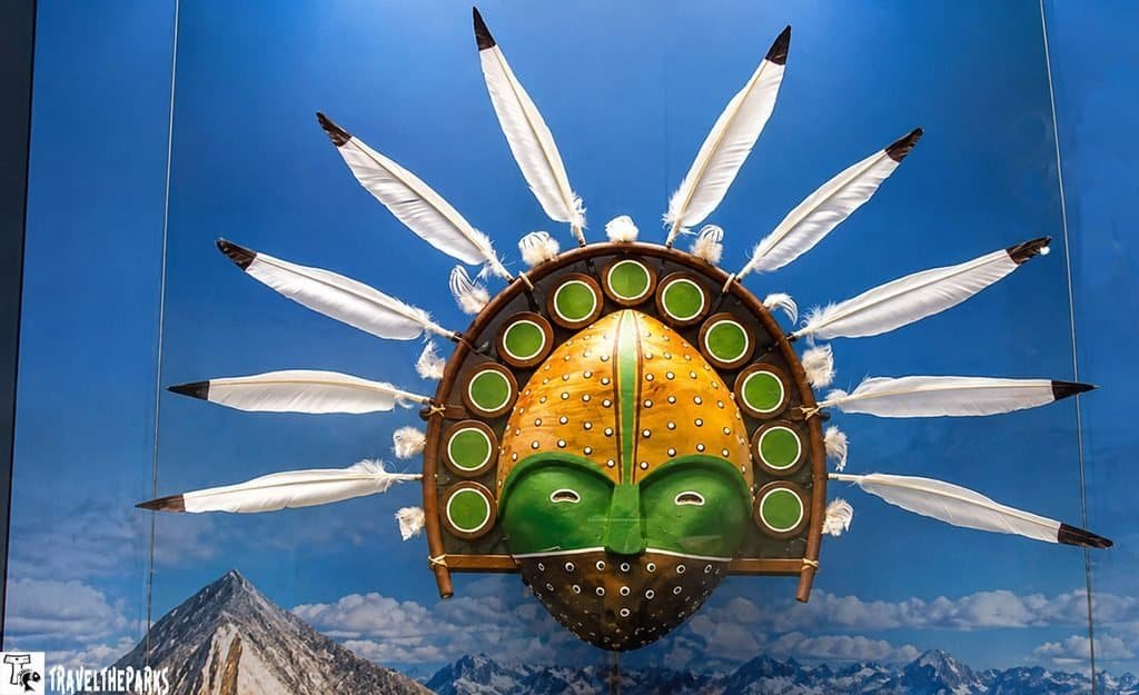 Colorful hot-air balloon shaped like a feathered mask floats against a blue sky, with a green-and-yellow face and mountain peaks below.
