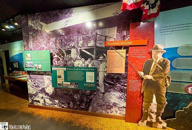 Museum exhibit with a large archival photo of a timber structure, interpretive panels, and a cutout of a man in a hat; the display has a historical tone.