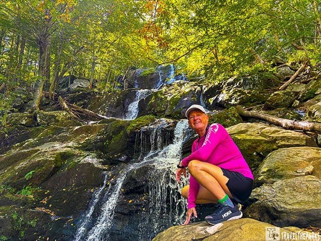 A smiling woman in a pink top and cap crouches on mossy rocks beside a cascading forest Dark Hallow Falls in Shenandoah NP, surrounded by sunlit autumn foliage.