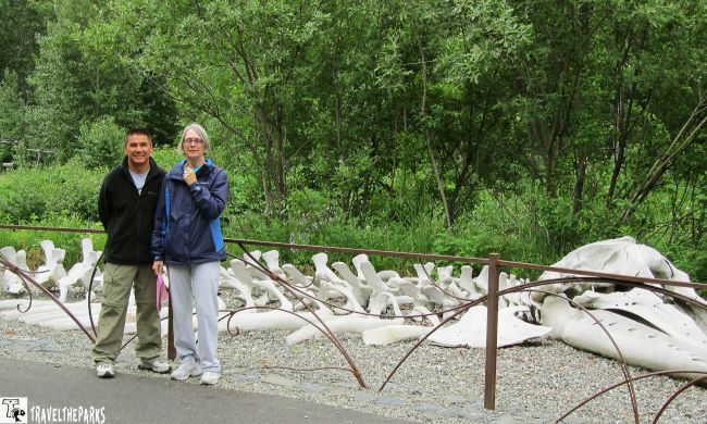 At the Alaska Heritage Center Village-Two people standing near large white bones, possibly whale bones, with a green foliage backdrop.