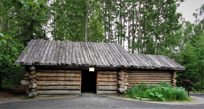 Athabaskan Village Lodge Traditional log cabin with a wooden roof in a forested area. At the Alaska Native Heritage Center