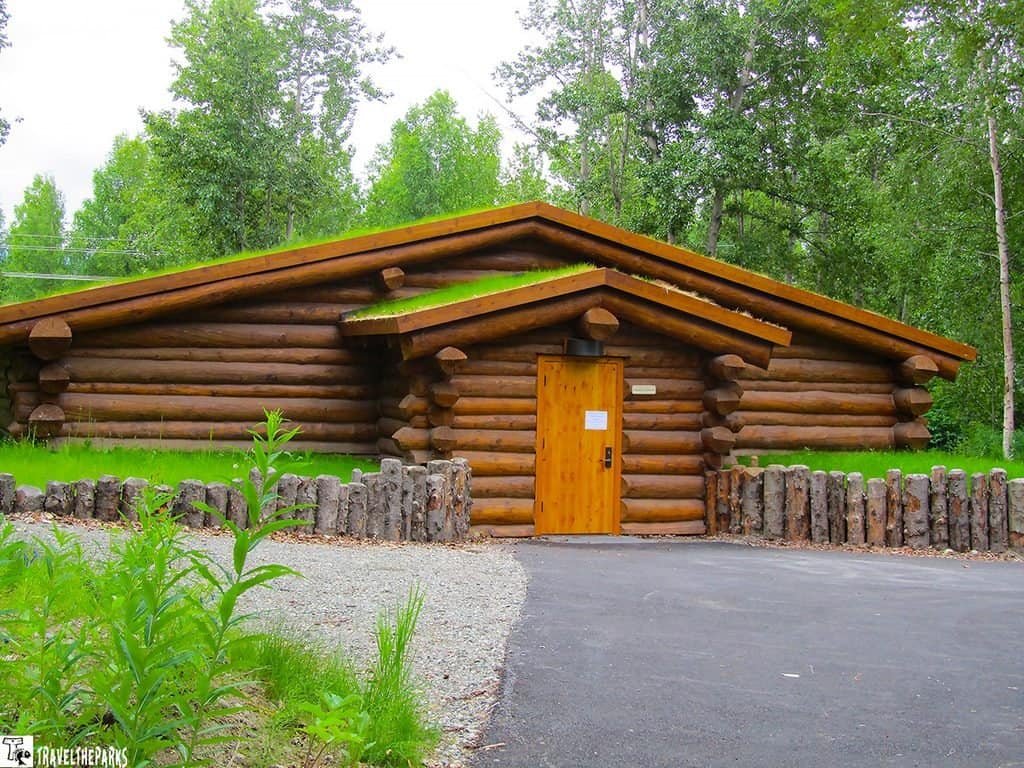 Rustic log cabin with layered wooden roof and bright door, set among lush green trees beside a gravel path and paved driveway; peaceful.