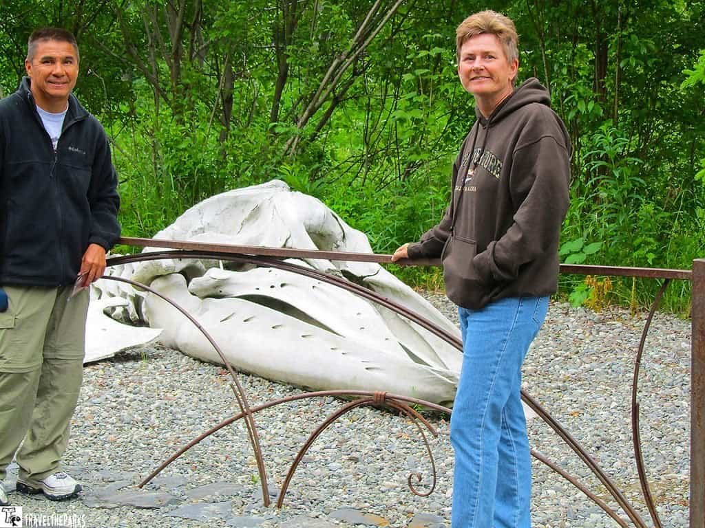 Two men stand beside a huge white whale sculpture in a wooded outdoor setting, smiling near a curved metal railing; peaceful, curious mood.