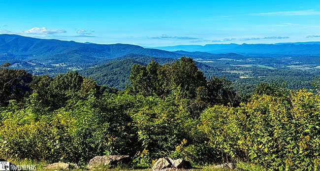 A sunny mountain overlook reveals dense green and yellow foliage, rolling blue ridges, and a broad valley beneath a clear sky, creating a peaceful, expansive mood.