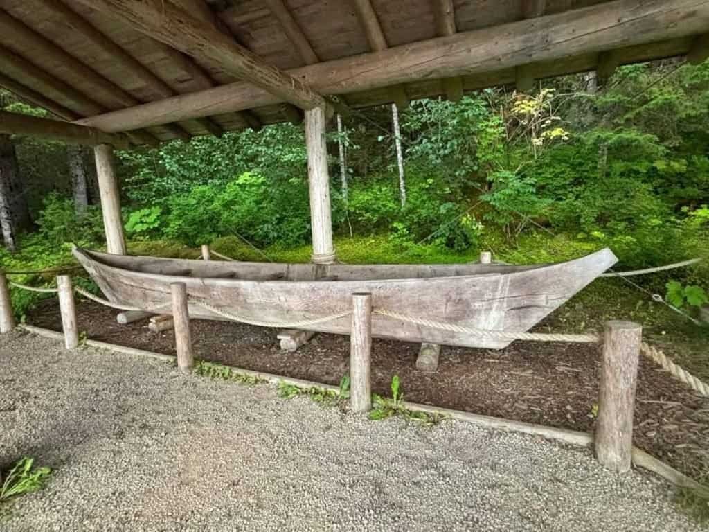 Weathered wooden canoe displayed beneath a rustic shelter in a lush green forest, behind a rope fence along a gravel path; serene setting.