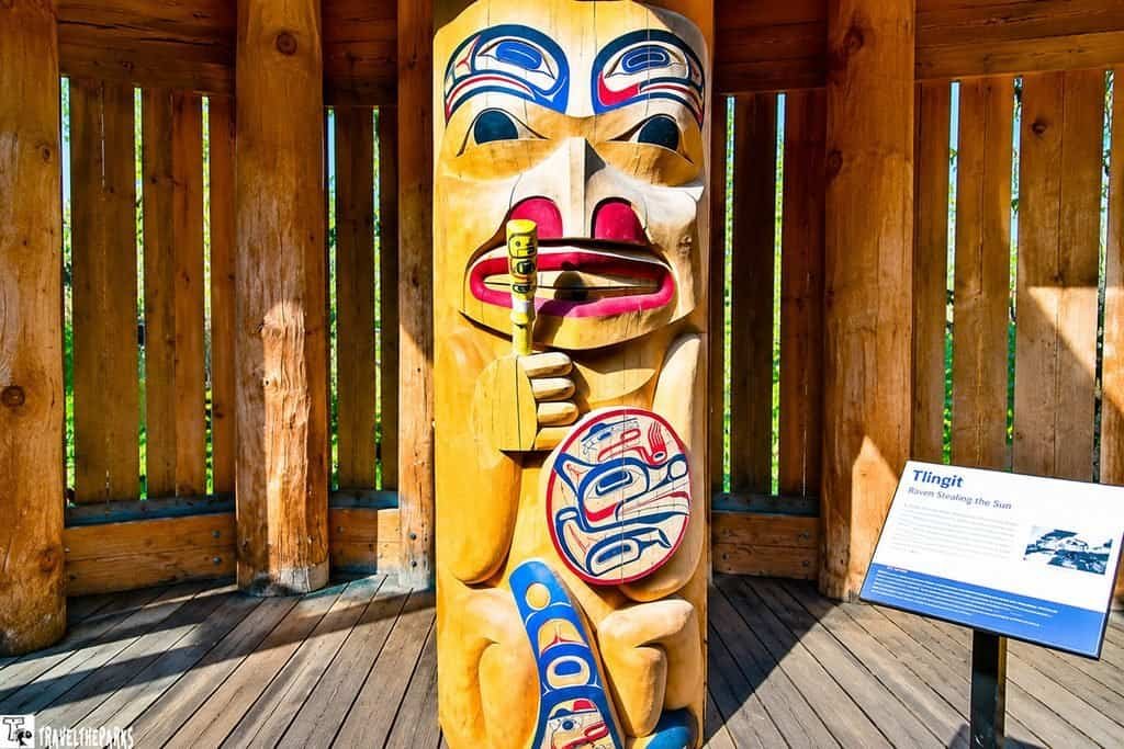 Colorful Northwest Coast Indigenous totem pole with a masked face and carved figures stands in a sunlit wooden shelter beside an information sign.