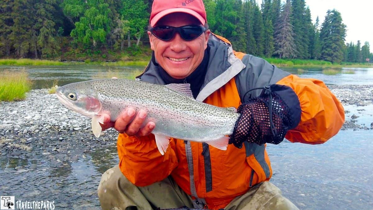 Person holding a large rainbow trout by the Kenai River, wearing outdoor gear.