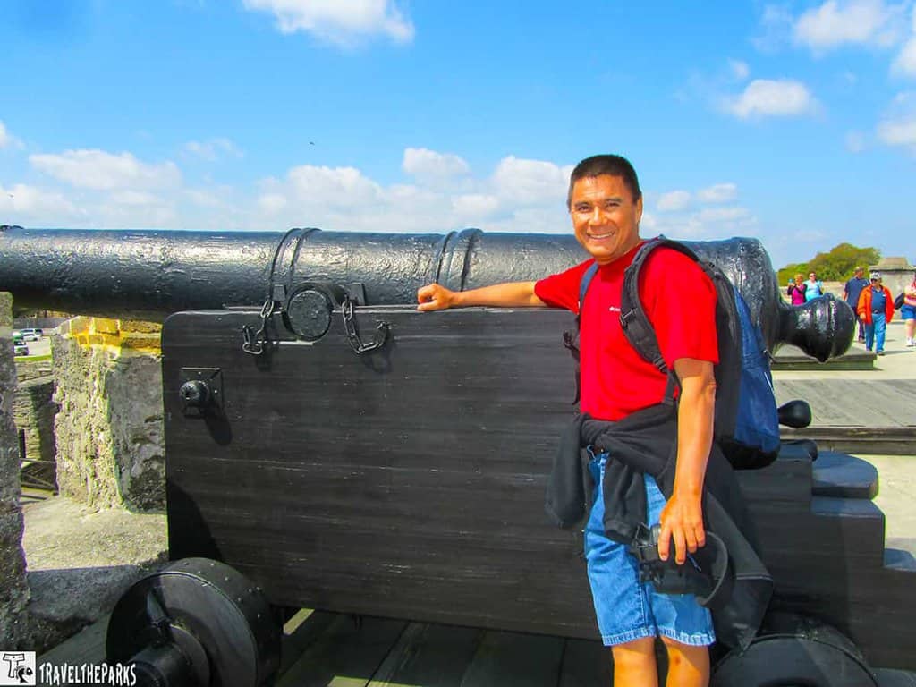 outdoor scene at a historic location. A person stands smiling next to a large, black, antique cannon mounted on a wooden carriage. The cannon and carriage are positioned on a stone fortification, likely part of a historic fort. The person is wearing a bright red shirt, blue shorts, and a backpack, with a jacket tied around their waist. The sky is bright blue with scattered white clouds, suggesting a sunny day. In the background, several people are seen walking on a wooden pathway, and some trees are visible in the distance.