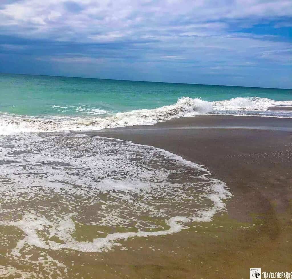 a serene beach scene at St. Augustine Beach. In the foreground, gentle waves wash over the wet, dark beige sand, creating a pattern of white foamy water. The middle ground features turquoise sea with small, cresting waves. Above, the sky is mostly cloudy, with patches of blue indicating clearer weather beyond. The scene is tranquil, emphasizing the blend of sea and sky.