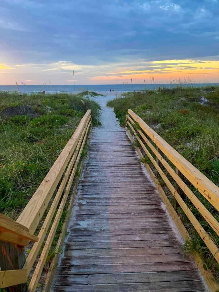 a wooden boardwalk leading towards a sandy beach, framed by wooden railings on either side. The boardwalk is slightly damp, possibly from recent rain or spray. On either side of the path, green vegetation with tall grass and a few sparse wildflowers grows. In the background, the beach meets a calm ocean under a vibrant sky that transitions from orange near the horizon to a soft blue higher up. The sky is filled with scattered clouds, illuminated by the setting or rising sun, casting a gentle glow over the scene.