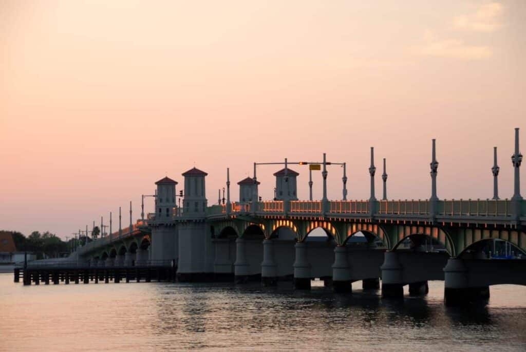the Bridge of Lions in St. Augustine, captured at sunset. The bridge extends across a calm body of water, with its series of arches reflected in the water below. The sky above is a gradient of soft pink and orange hues, indicative of the setting sun. Along the bridge, there are multiple towers, each with a red-tiled roof, evenly spaced along its length. Decorative light posts and railings are visible, casting a warm glow. The foreground showcases the bridge piers, and the background includes a few trees and buildings.