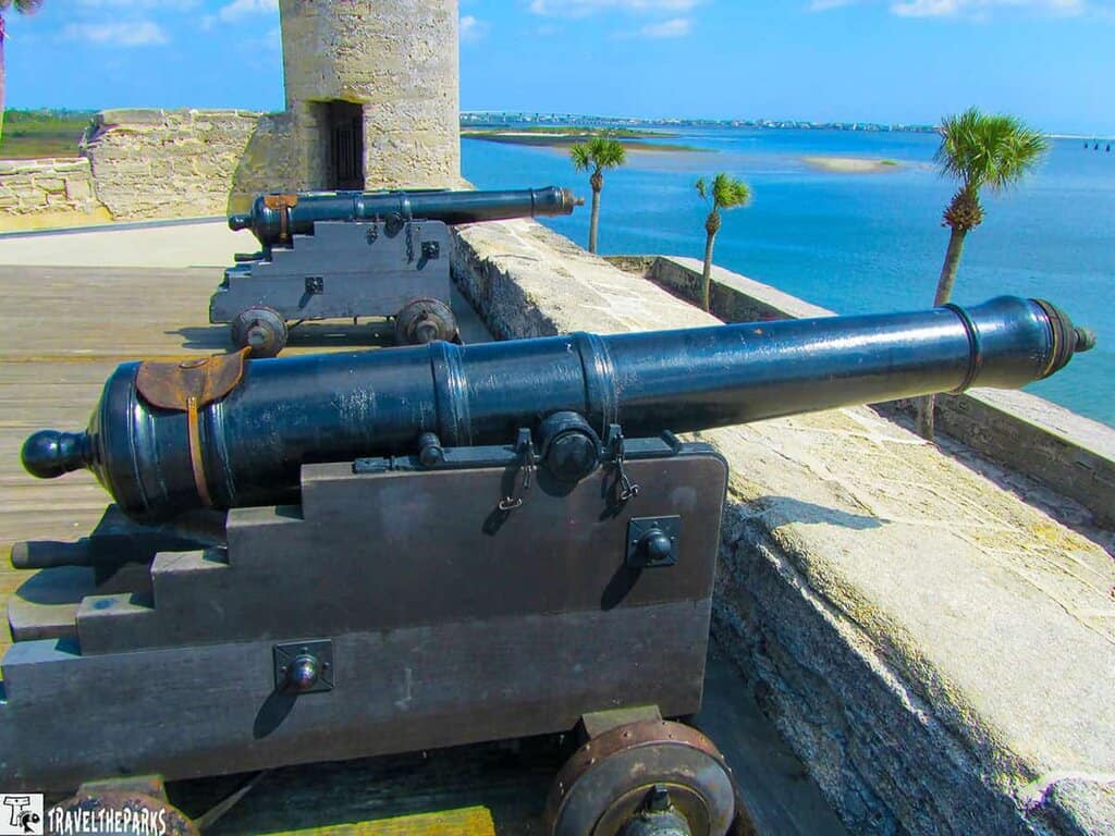 a historical scene at the Castillo de San Marcos, with two large black cannons mounted on wooden carriages atop a stone fort wall. The foreground cannon is prominently displayed, showcasing its metal surface and intricate detail. The stone wall extends into the distance, separating the cannons from a scenic view of a body of water. In the background, several palm trees are dispersed along the coastline, and a broad expanse of blue water stretches towards the horizon under a partly cloudy sky.