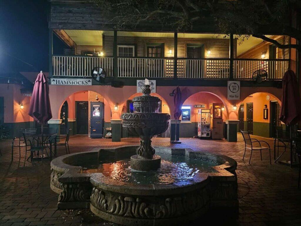 a courtyard at night in St. Augustine, with a prominent stone fountain in the foreground. The fountain has intricate carvings and a small pool of water at its base, reflecting nearby lights. Surrounding the fountain are several metal chairs and tables, some under large burgundy umbrellas. In the background, a two-story building features a row of arched doorways and windows on the ground level, with a covered balcony on the second floor. The building's exterior is painted in warm tones of orange and green. Various lights illuminate the courtyard and building, creating a cozy atmosphere.